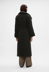 Casaco longo de cor verde escura com uma cintura cingida, colarinho largo e punhos com botões. Feito de um material espesso, tem um corte reto e sem padrões visíveis.