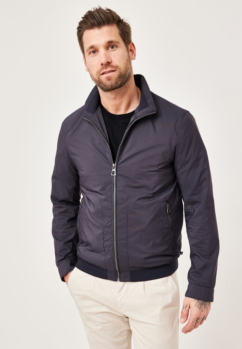 Navy striped bomber jacket with a high collar and zip closure, featuring side pockets and ribbed cuffs, paired with light-colored trousers.