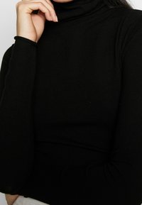 Person som bär en åtsittande svart polotröja, vilar en hand nära hakan mot en enkel vit bakgrund.