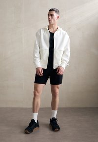 Sweat à capuche blanc zippé avec poches avant, débardeur noir et shorts noirs. Chaussures noires avec semelles en gomme et chaussettes blanches. Design simple et moderne.