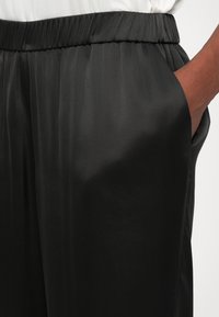 Roka roka melnajās satīna biksēs ar elastīgu jostasvietu, valkāts ar baltu krekla, parādot auduma tekstūru un brīvu piegriezumu.