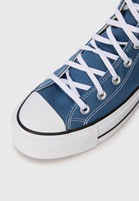 Blå kanvas högbent sneakers med vita snören, gummikappa och texturerad vit sula. Har silverfärgade öglor och svarta accentlinjer.