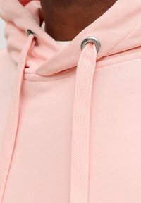 Felpa con cappuccio rosa caratterizzata da un tessuto strutturato, scollo tondo e occhielli metallici per i cordoncini. Vista ravvicinata del cappuccio e delle cuciture.