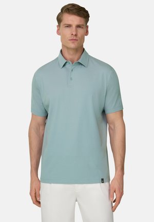 Mann trägt ein hellblaues Polohemd mit kurzen Ärmeln und weiße Hosen, steht vor einem einfarbigen hellen Hintergrund.