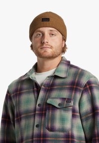 Bruine geribbelde gebreide beanie met een zwart logo-label, gedragen met een meerkleurig geruit flanellen overhemd met groene, paarse en oranje tinten.