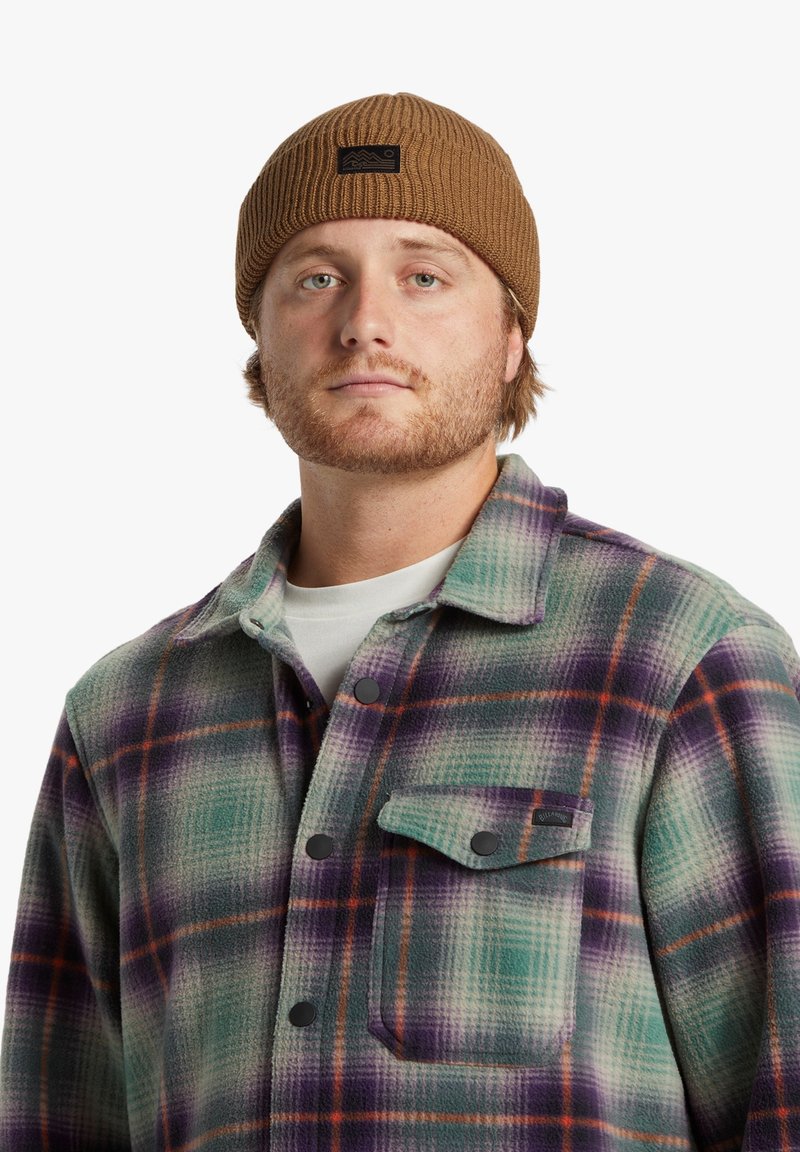 Bonnet en tricot côtelé marron avec une étiquette logo noire, porté avec une chemise en flanelle à carreaux multicolores comportant des tons verts, violets et oranges.