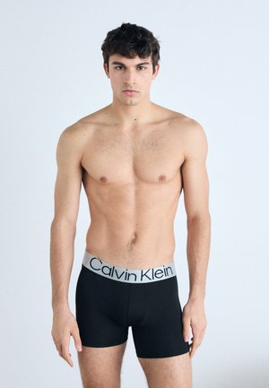 Črne bokserice z grivno elastično pasom, na katerem je napis "Calvin Klein" v črni barvi. Model stoji z nevtralnim izrazom.
