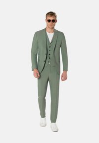 Hellgrüner Anzug mit strukturierter Oberfläche, bestehend aus einem taillierten Blazer, einer Weste mit drei Knöpfen und schmal geschnittenen Hosen, kombiniert mit weißen Sneakers.