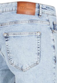 Jeans en denim bleu clair avec une patch en cuir à la taille, présentant une poche arrière avec un contour brodé et une texture froissée.