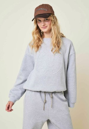 Jeune femme aux cheveux blonds ondulés portant une casquette marron et un sweat-shirt gris clair assorti avec un pantalon de survêtement à cordon.