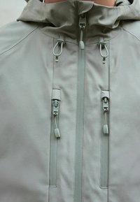 Veste gris clair avec fermeture éclair frontale, deux poches poitrine verticales zippées, et cordons de capuche réglables près du cou.