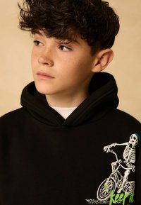 Un garçon avec des cheveux noirs et bouclés et des taches de rousseur portant un sweat à capuche noir avec un graphique de squelette faisant du vélo sur le devant.
