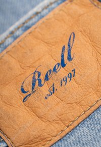 Etiqueta de cuero con texto azul que dice "Reell est. 1997" cosida sobre tela de mezclilla, con una superficie de tono marrón texturizada y costuras visibles.