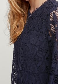 Blouse en dentelle bleu marine avec des motifs floraux complexes, des manches semi-translucides et un col à festons. Tissu léger avec une texture lisse.