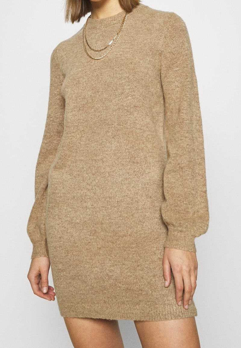 Beige neulemekko, jossa on pitkät puffi hihat ja pyöreä pääntie. Pehmeä tekstuuri, minimalistinen muotoilu ja helma juuri polven yläpuolella.