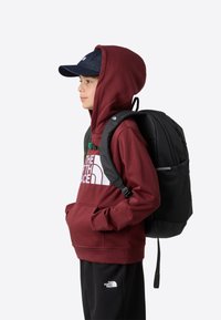 Burgunderfarget hettegenser med en logo, svart ryggsekk med polstrede stropper, svarte bukser med en liten logo, og en mørk denimcaps.