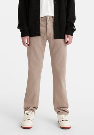 Persoon draagt beige rechte broek, witte sneakers met rode zolen, wit overhemd en zwarte gebuttonneerde cardigan, getoond van midden torso naar beneden.