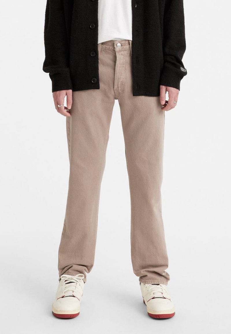 Personne portant un pantalon beige droit, des baskets blanches à semelles rouges, une chemise blanche et un cardigan noir boutonné, vue de la moitié du torse vers le bas.