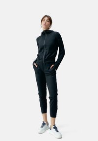 Completo sportivo nero con giacca zip e pantaloni affusolati, realizzato in un tessuto liscio, abbinato a sneaker bianche e polsini elasticizzati.