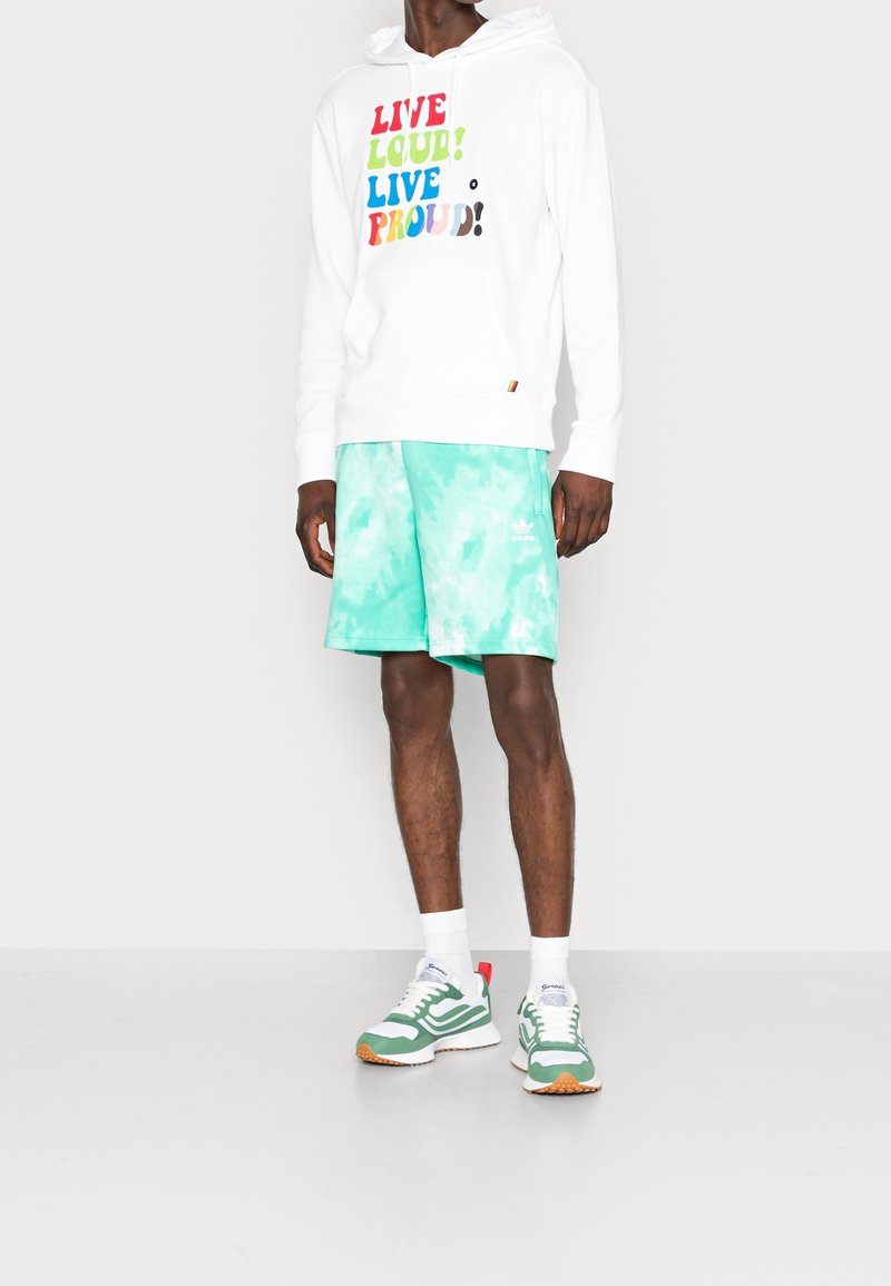 Person som bär en vit huvtröja med färggivande texten "Live Loud! Live Proud!", mintgröna tie-dye shorts, vita strumpor och gröna sneakers.