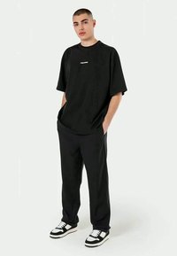 Tricou oversized negru cu un mic logo alb pe piept, asortat cu pantaloni negri și adidași black and white, pe un fundal neutru.