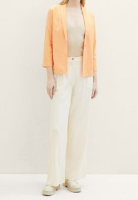 Blazer orange léger, haut beige ajusté et pantalon large crème. Texture lisse, un seul bouton sur le pantalon, chaussures décontractées.