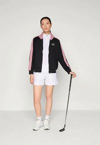 Juoda ir rožinė sportinė striukė su užtraukiama apykakle, balta polo marškinėliai ir balti šortai; stovi su golfo lazda. Sneakeriai užbaigia aprangą.