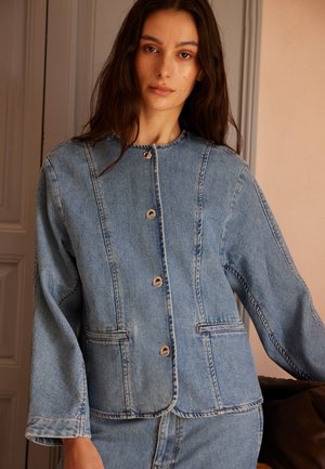 Kvinne med langt mørkt hår iført en lyseblå denimjakke med sølvknapper, stående innendørs mot en beige vegg og dørkarm.