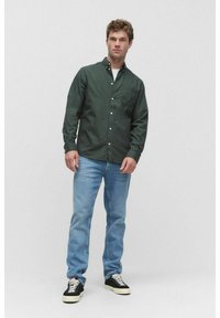 Man in een donkergroene blouse, lichtblauwe jeans en zwarte sneakers die tegen een effen lichte achtergrond staat.