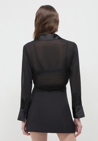 Frau mit welligem braunen Haar trägt eine durchsichtige schwarze Bluse mit langen Ärmeln und einen schwarzen Rock, von hinten vor einem schlichten Hintergrund gezeigt.