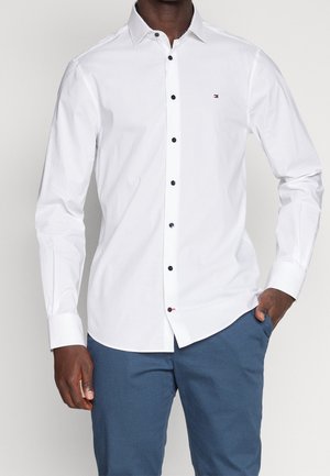 Man wearing white long-sleeve button-up shirt with black buttons and small flag logo, paired with blue pants, hand in pocket.