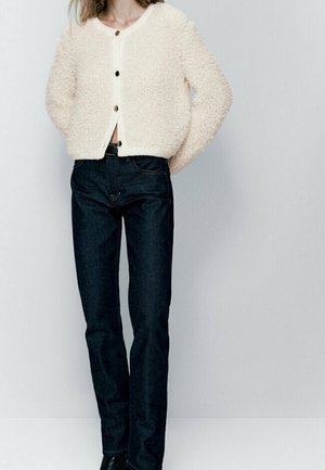 Vrouw die een crèmekleurige, pluizige, knoopvest draagt en hoge taille, donkerblauwe jeans met rechte pijpen, tegen een effen achtergrond.