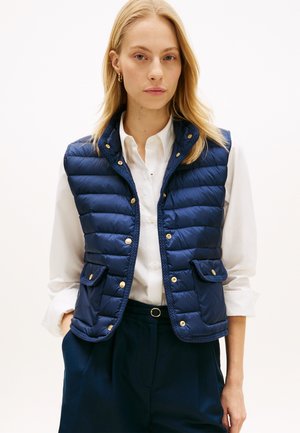 Tmavomodrá prešívaná vesta s patentnými gombíkmi, dvoma prednými vreckami s klopami a golierom. Nosená cez bielu košeľu, so hladkou textúrou.