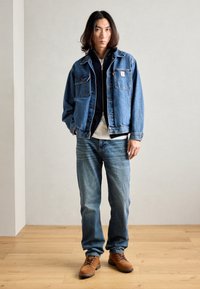 Chaqueta de denim en azul medio con dos bolsillos frontales y costuras blancas, combinada con jeans de color azul claro y botas marrones con cordones sobre un suelo de madera.