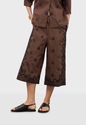 Pantaloni marroni a gamba larga e taglio corto con motivi floreali neri ricamati, abbinati a sandali neri aperti e top marrone coordinato.