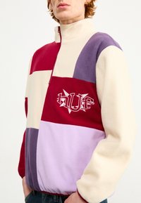 Farveopdelt fleecejakke med lynlås i rød, creme, lavendel og lilla med broderet "HUF" logo. Blød tekstur og afslappet pasform.