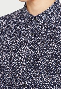 Námornícká košile s knoflíky s malým květinovým vzorem v růžové a modré barvě, klasickým límcem a černými knoflíky. Hladká textilní struktura.