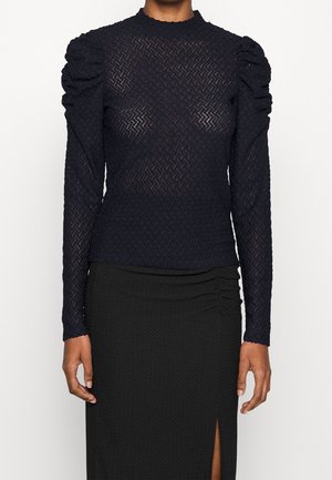 Kobieta ubrana w czarną, teksturowaną bluzkę z długim rękawem, z marszczonymi ramionami oraz czarną spódnicę z przednim rozcięciem.