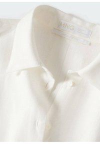 Chemise en lin blanc à boutons avec un col classique, présentant une texture subtile et une étiquette indiquant "MNG Édition Limitée."