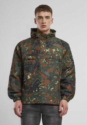 Mand iført en grøn, brun og sort camouflage hættetrøje med elastiske manchetter og en foran lynlåslomme, stående mod en grå baggrund.