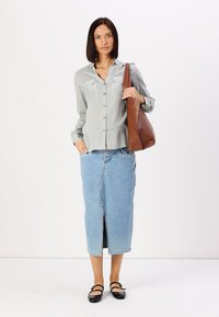 Camicia a righe con due tasche sul petto, abbinata a una gonna di jeans azzurri chiari con uno spacco frontale. Borsa a spalla marrone e ballerine nere.