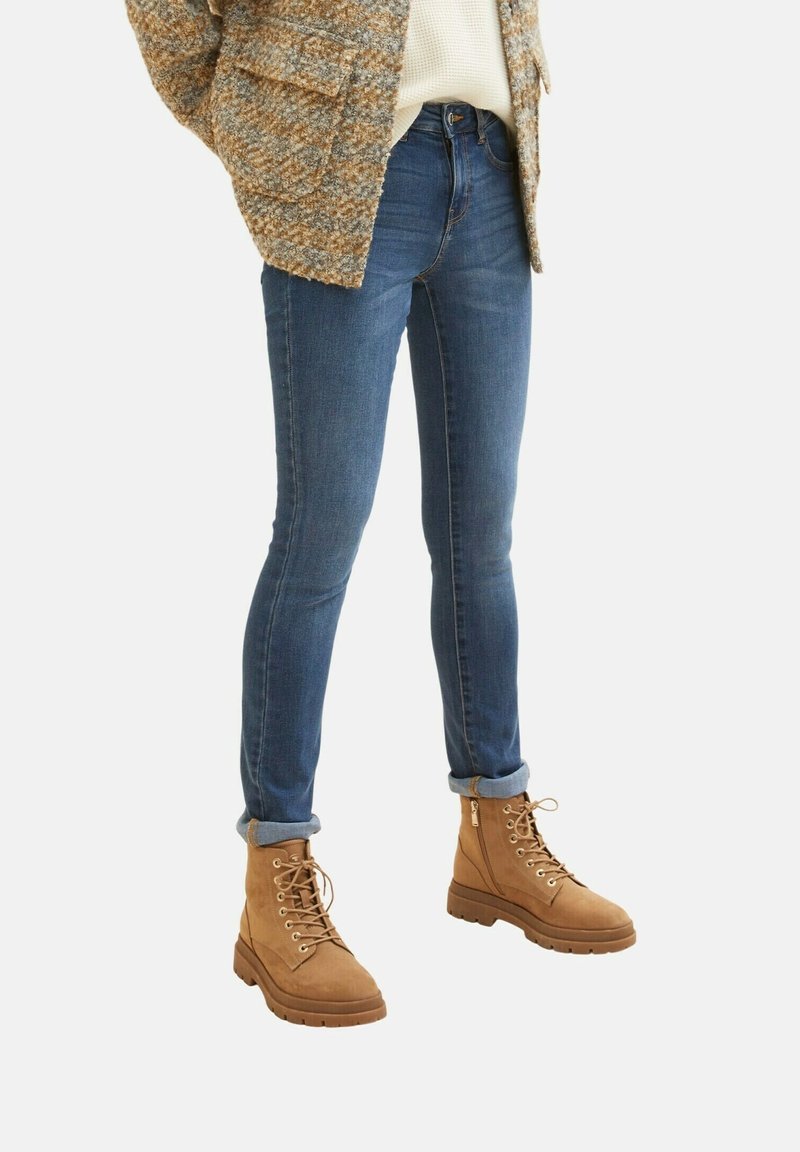 Modré džíny skinny s středně vysokým pasem, v kombinaci s hnědými šněrovacími kotníkovými botami s robustní podrážkou a ohrnutými manžetami.