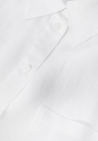 Camisa blanca de cuello con botones visibles y bolsillo en el pecho, mostrando una textura de tela suave y detalles de costura impecables.