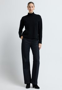 Kobieta stoi z rękami w kieszeniach, ubrana w czarny sweter z długim rękawem, ciemnoniebieskie jeansy, czarne botki na obcasie i złote kolczyki w kształcie kółek.