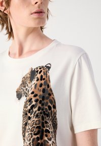 T-shirt blanc avec un graphisme de léopard sur le côté, présentant des taches noires détaillées et une texture en coton lisse.