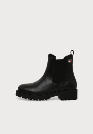 Bottine noire en cuir avec panneau latéral élastique, semelle épaisse, languette de tirage et petite étiquette logo près du haut sur un fond uni.