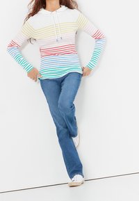 Haut à capuche à manches longues avec des rayures horizontales multicolores, principalement blanches, jaunes, roses, rouges, bleues et vertes ; assorti à un jean bleu et des chaussures blanches.