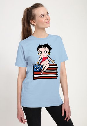 Lichtblauw t-shirt met een grafisch ontwerp van een cartoonfiguur die op een Amerikaanse vlag zit, met een hart en gouden hoepeloorbellen.
