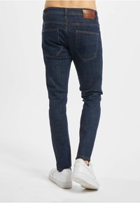 Mørkeblå slanke jeans med glatt tekstur, med baklommer og en brun skinnlap ved midjebåndet. Hvite joggesko synlige under.