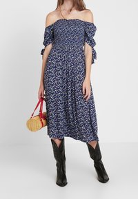 Off-shoulder blå klänning med blommönster, smockad midja, korta volangärmar och svängig kjol; kombinerad med svarta cowboyboots och en rund väska.